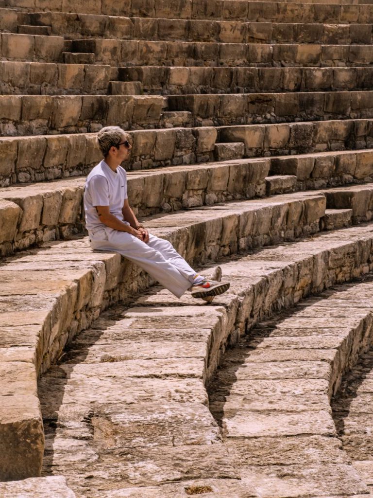 Un giovane seduto sui gradoni di pietra di un antico teatro greco-romano (probabilmente Kourion). È di profilo, indossa abiti chiari e guarda verso l'orchestra, circondato dalla maestosa struttura semicircolare in pietra baciata dal sole