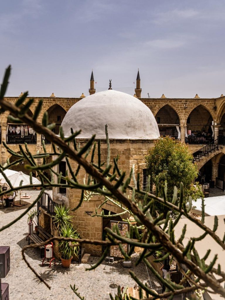 Una prospettiva dall'alto del cortile interno del Büyük Han a Nicosia durante un viaggio fly&drive a Cipro. In primo piano dei rami verdi sfocati incorniciano la cupola centrale in pietra e le arcate a sesto acuto del piano superiore, con i minareti che svettano sullo sfondo
