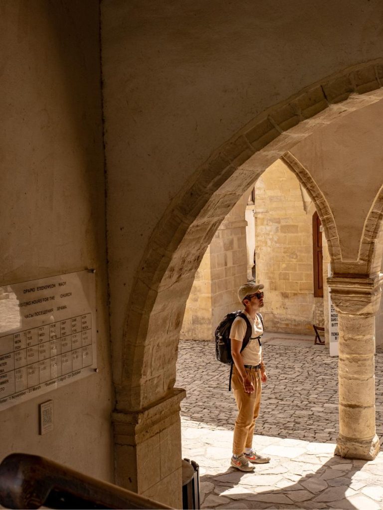 Un ragazzo con zaino e cappellino osserva un cortile storico attraverso un antico arco in pietra. La luce del sole illumina il pavimento in ciottoli, creando un forte contrasto con l'ombra dell'arcata in primo piano