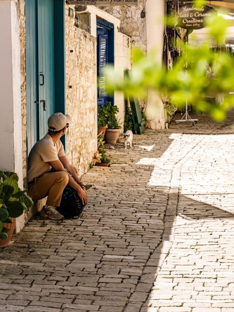 Uno scorcio di un tipico vicolo cipriota con pareti in pietra chiara e porte blu. Un ragazzo è seduto su uno scalino in ombra sulla sinistra, mentre un gatto bianco e tigrato cammina sulla strada lastricata in lontananza